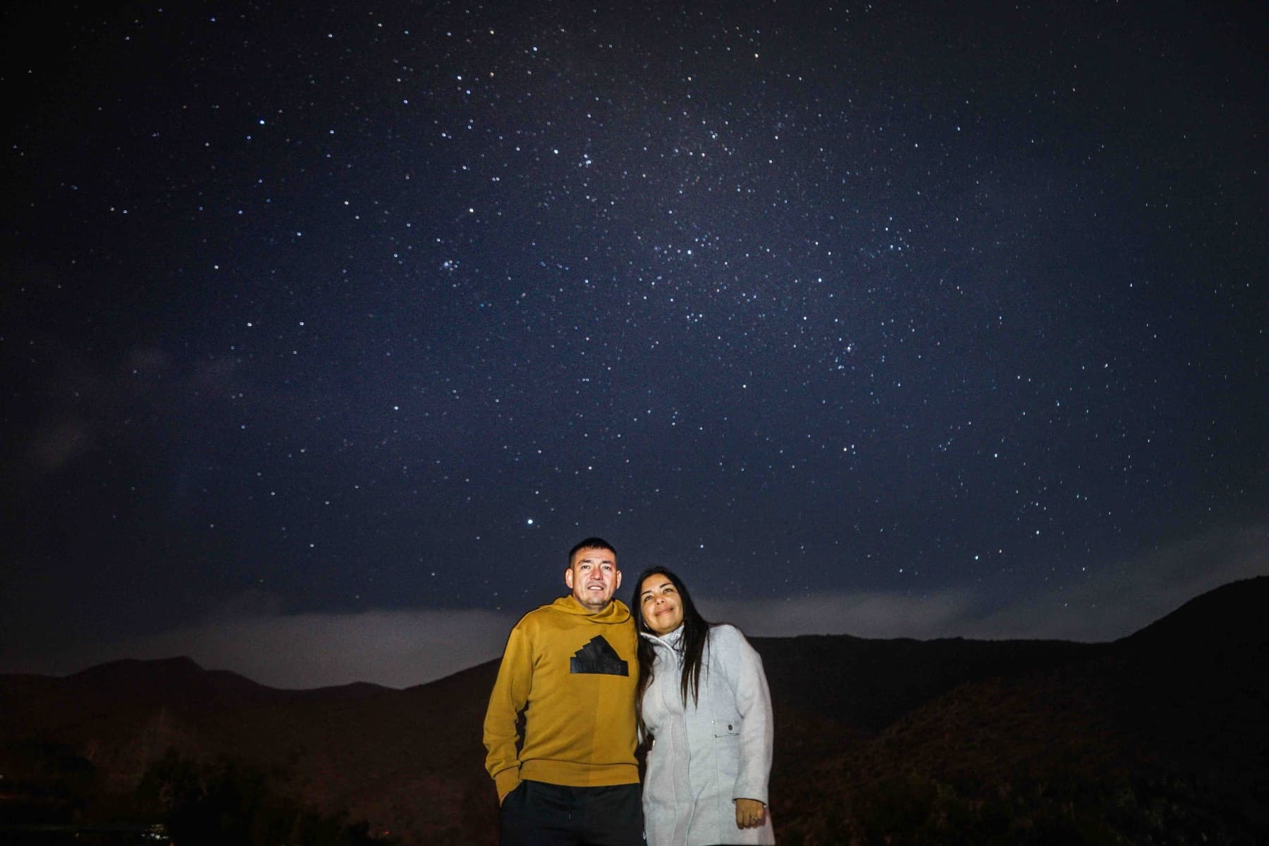 Una pareja de visitantes posa bajo el cielo estrellado del Molle, con los cerros del valle al fondo.