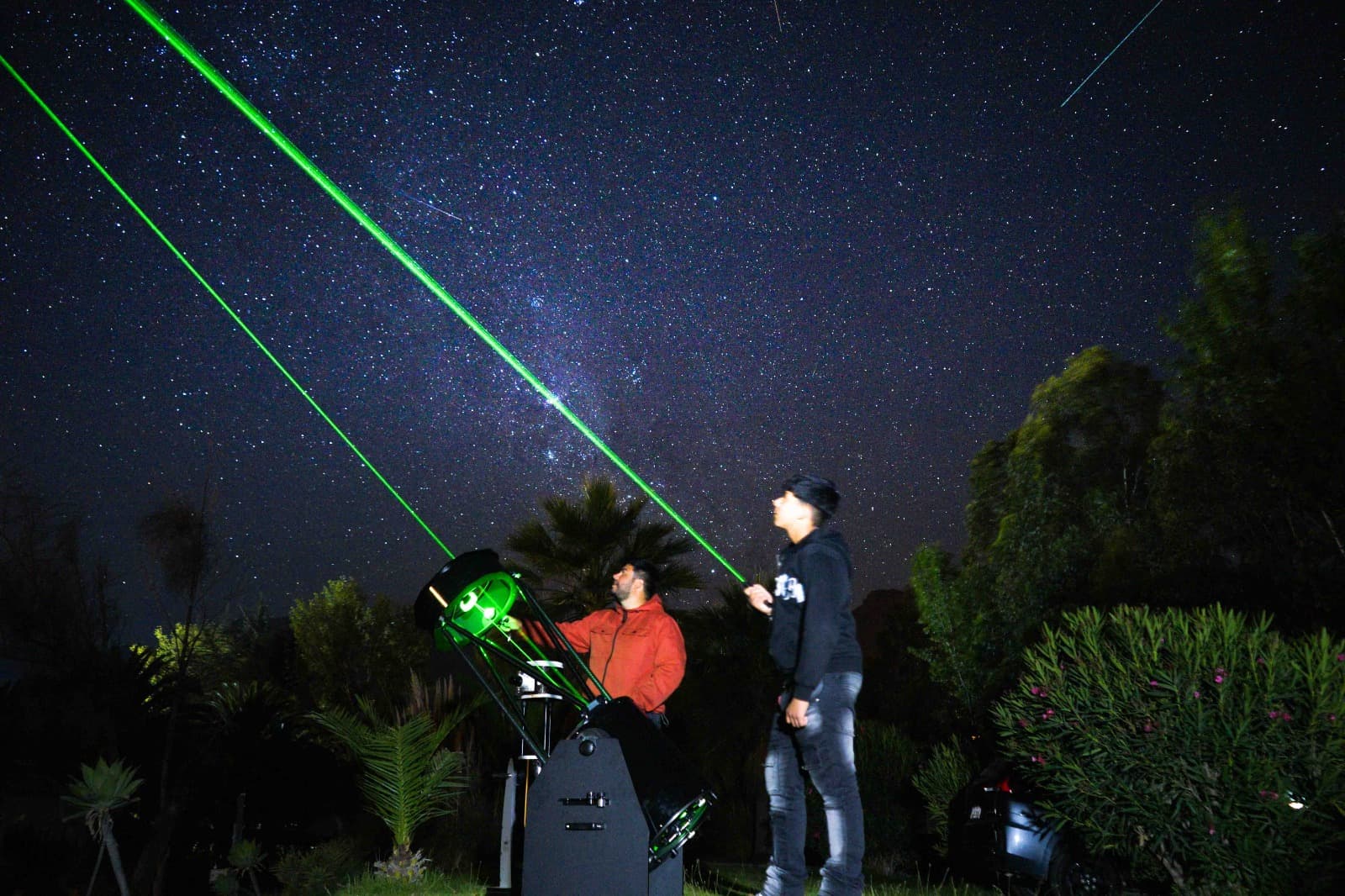 Nuestro guía junto a un gran telescopio y un visitante bajo la Vía Láctea, mientras dos láseres verdes señalan el cielo en El Molle.